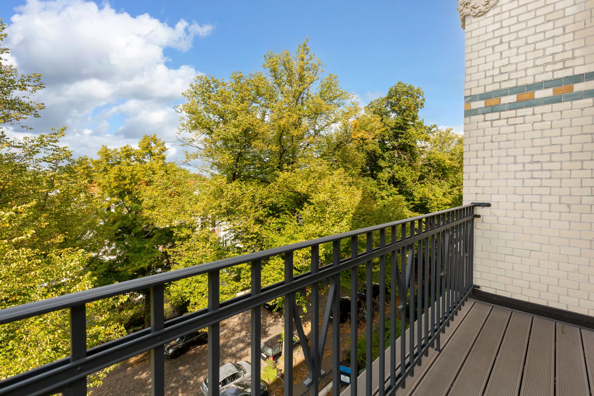 Balkon mit schwarzem Geländer, Blick auf grüne Bäume und einige geparkte Autos darunter.