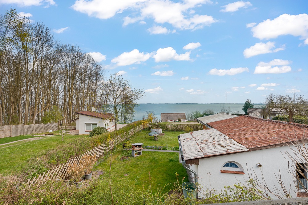 Garten mit Rasen, Bäumen, kleinem Haus und Blick auf einen See im Hintergrund.