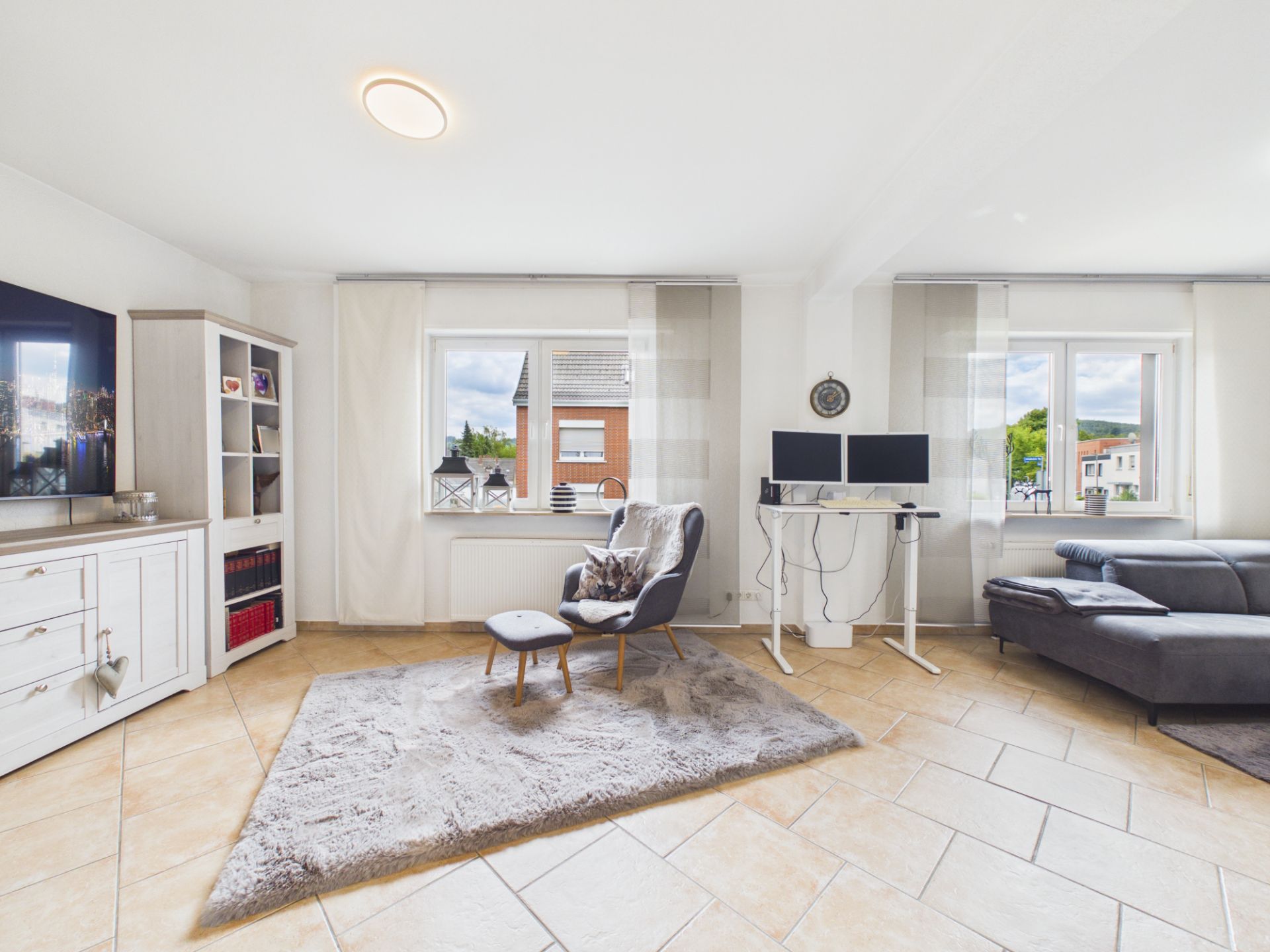 Helles Wohnzimmer mit Sessel, Teppich, Ecksofa und Schreibtisch mit zwei Monitoren.