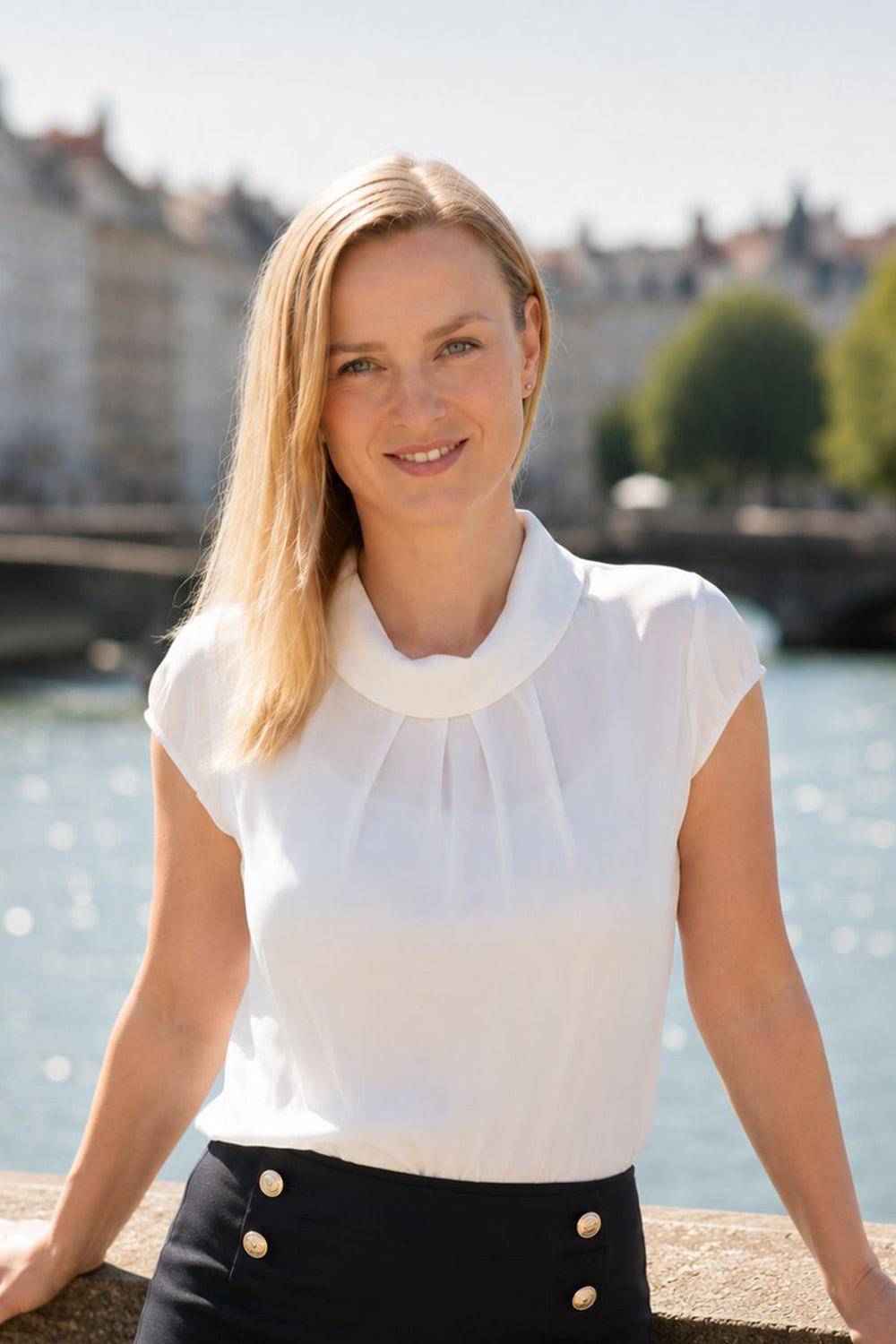 Frau mit blondem Haar und weißer Bluse steht vor Wasser mit Booten im Hintergrund.