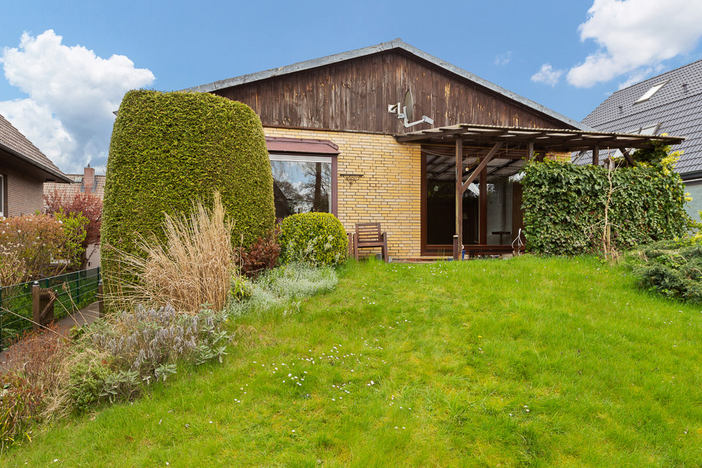 Backsteinhaus mit großer Fensterfront, Terrasse, Rasen und dichtem Strauch im Garten.