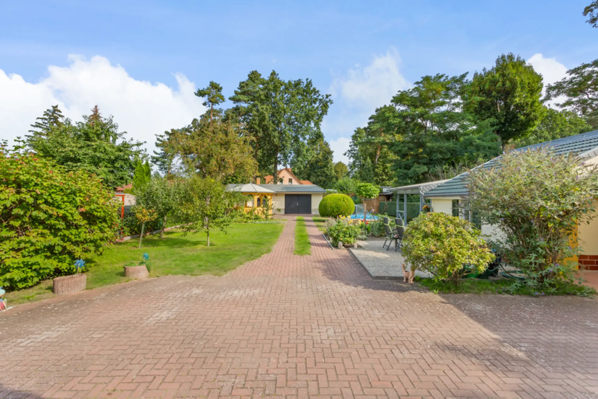 Gepflasterte Einfahrt mit Garten, Bäumen und Garage im Hintergrund.