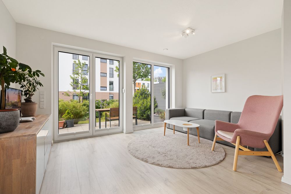 Helles Wohnzimmer mit grauem Sofa, rosa Sessel, Teppich und großen Fenstern zum Garten.