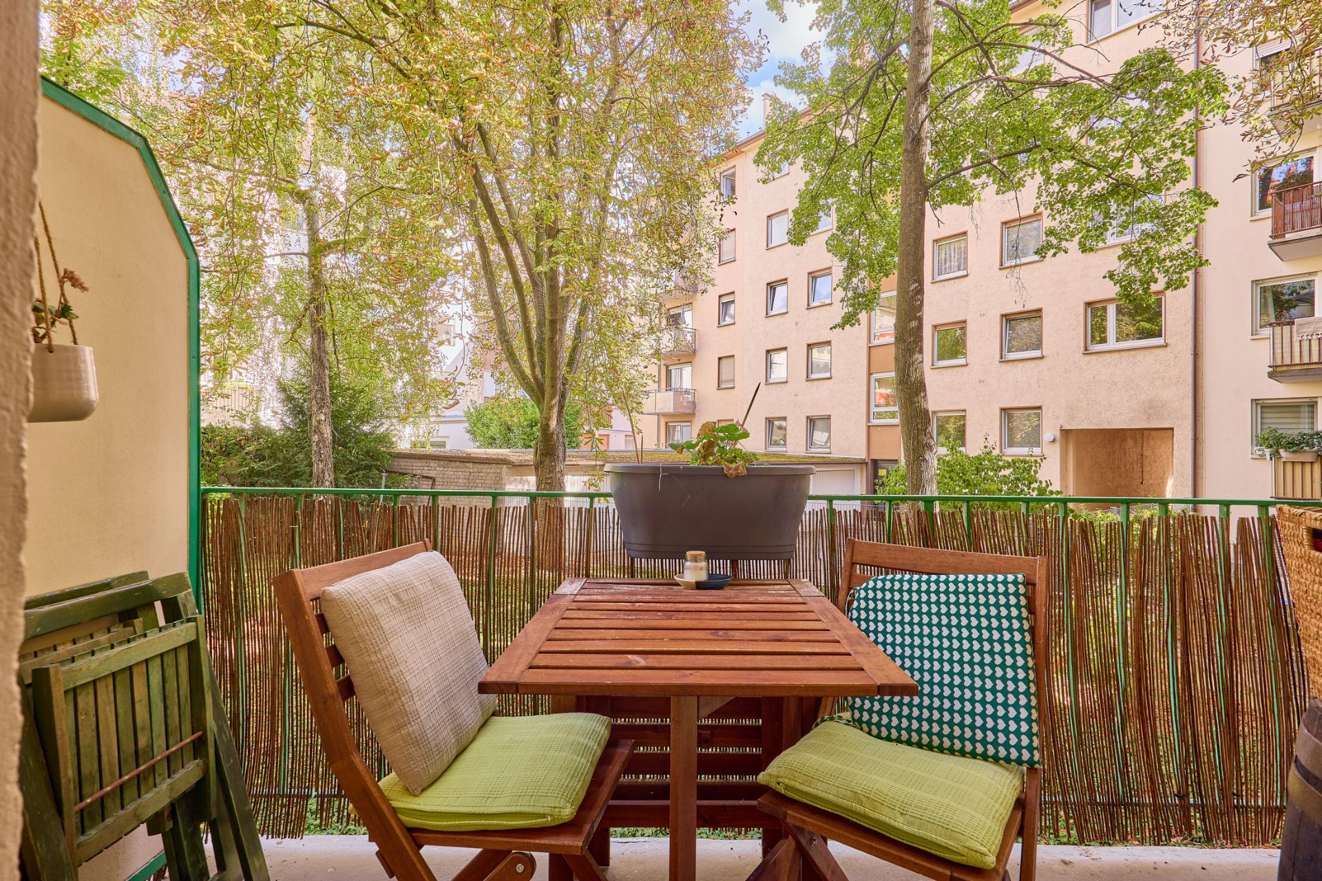 Balkon mit Holztisch und zwei Stühlen, Blick auf Bäume und Wohngebäude im Hintergrund.