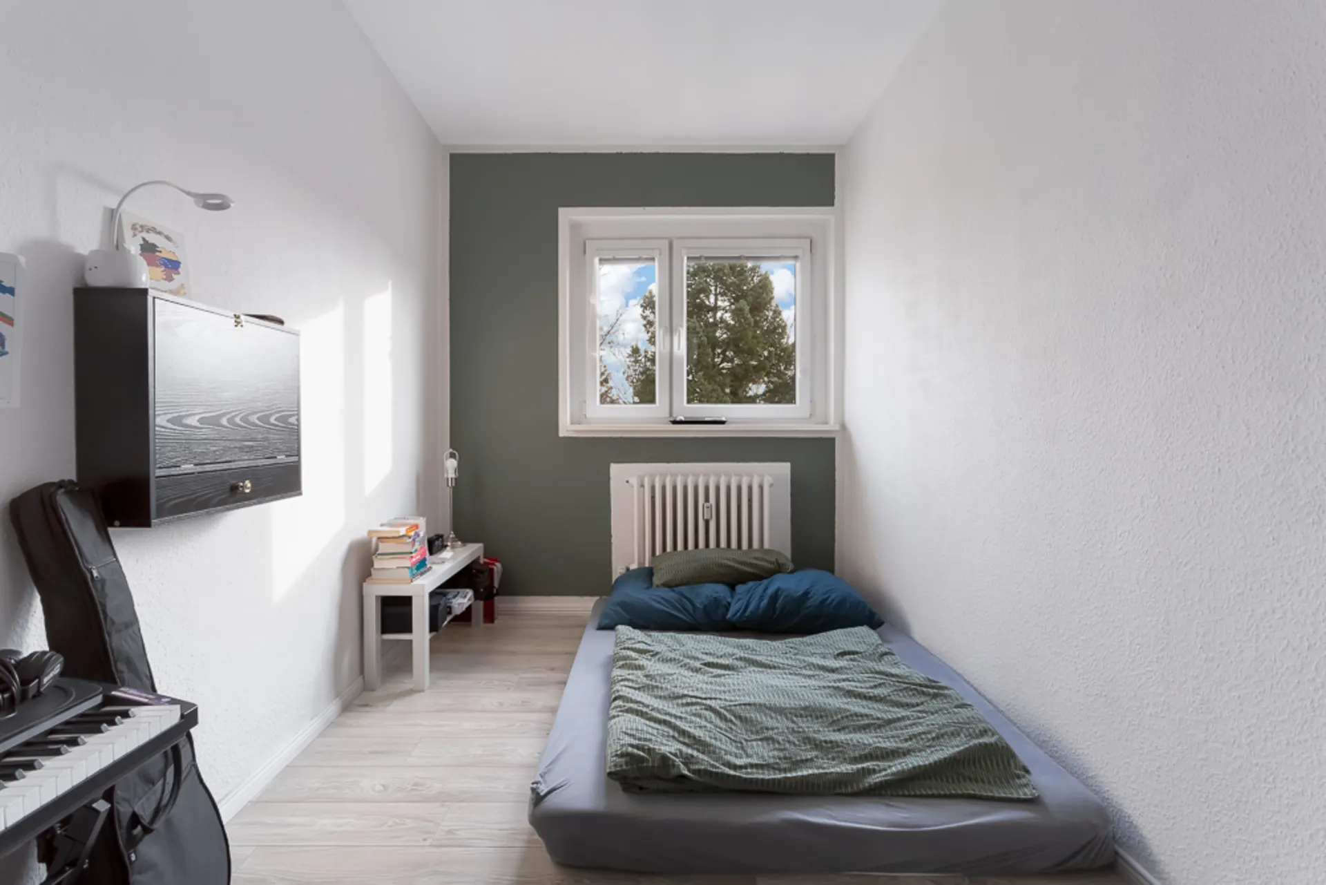 Schmales Schlafzimmer mit Matratze, Tisch, Büchern und Fenster zur Außenseite.