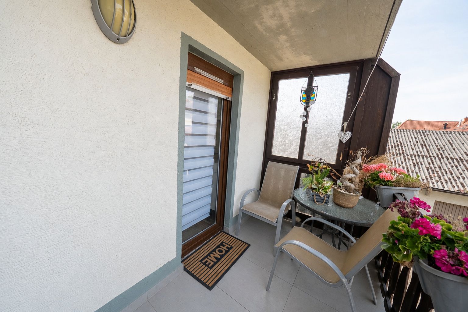 Kleiner Balkon mit zwei Stühlen, Tisch, Blumen und einer Türmatte vor der Tür.