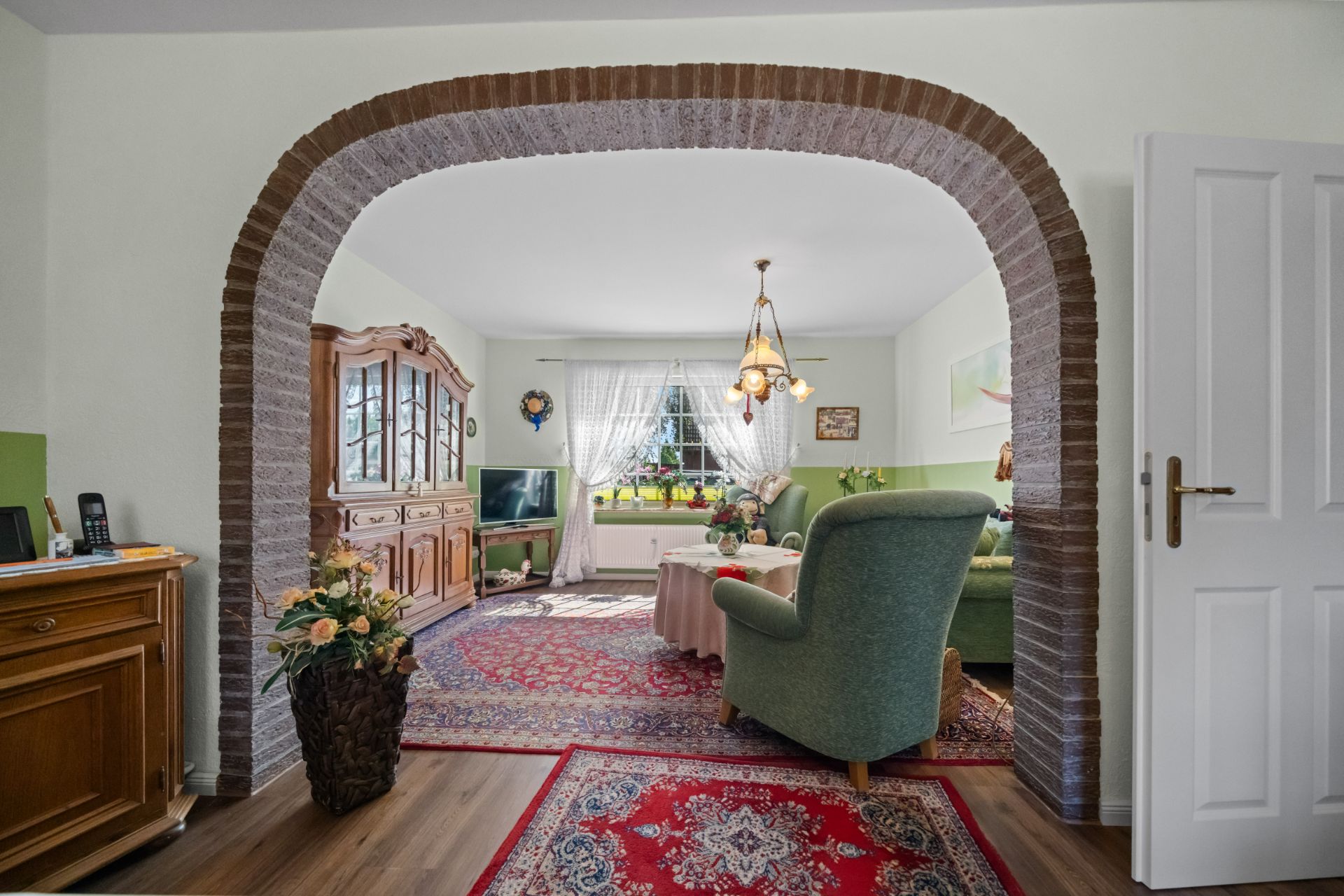 Wohnzimmer mit Teppichen, grünen Polstermöbeln, Holzmöbeln und gemauertem Rundbogen.