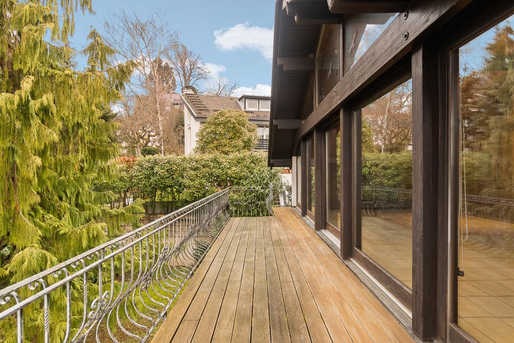 Holzterrasse mit Geländer an einem Wohnhaus, umgeben von Bäumen und großen Fenstern.
