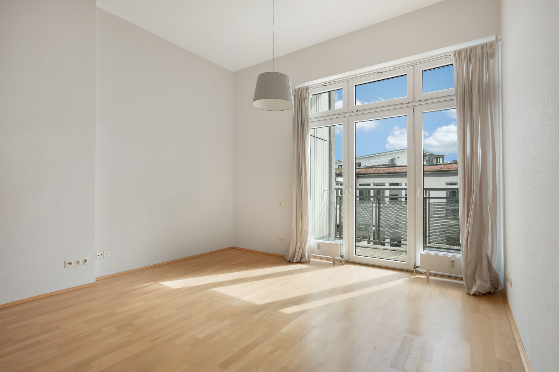 Helles, leeres Zimmer mit Holzfußboden, großen Fenstern und Vorhängen.