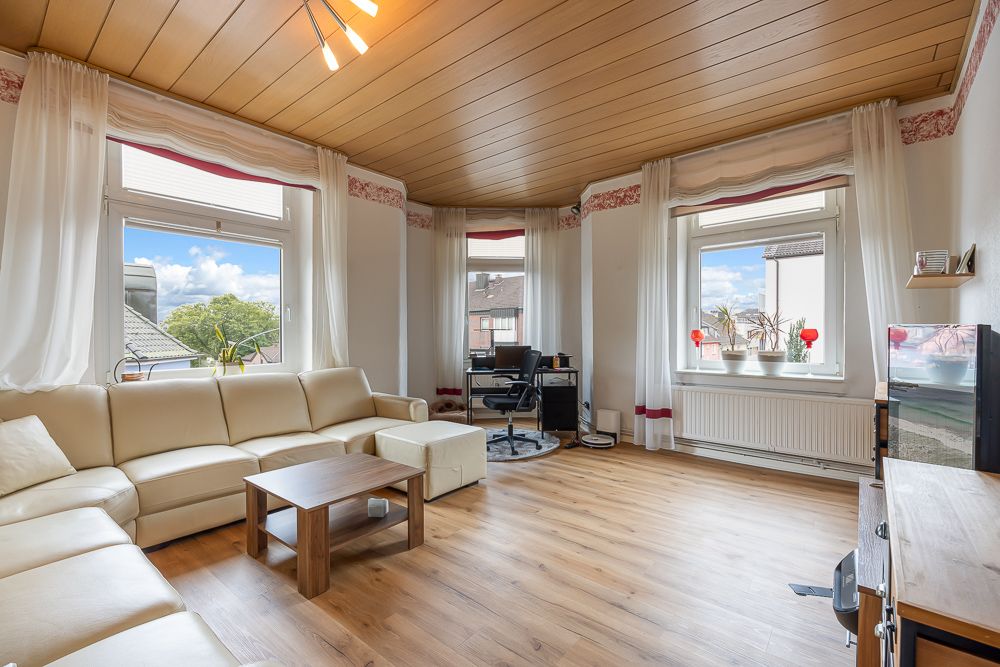 Helles Wohnzimmer mit Ecksofa, Couchtisch, Schreibtisch und großen Fenstern mit weißen Vorhängen.