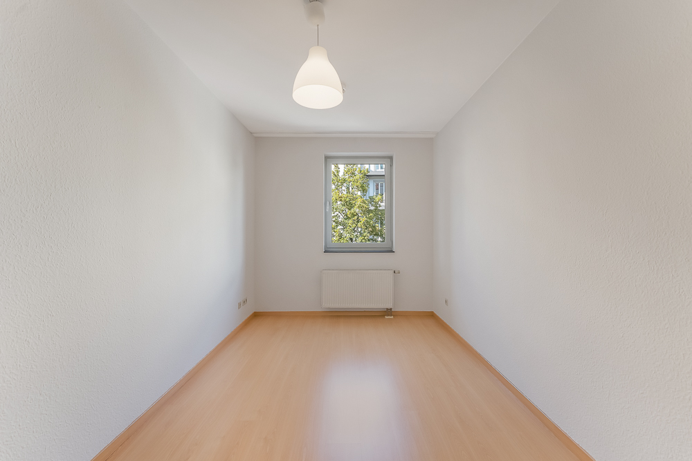Leerer, heller Raum mit Holzboden, weißer Decke, Fenster, Heizung und Deckenlampe.