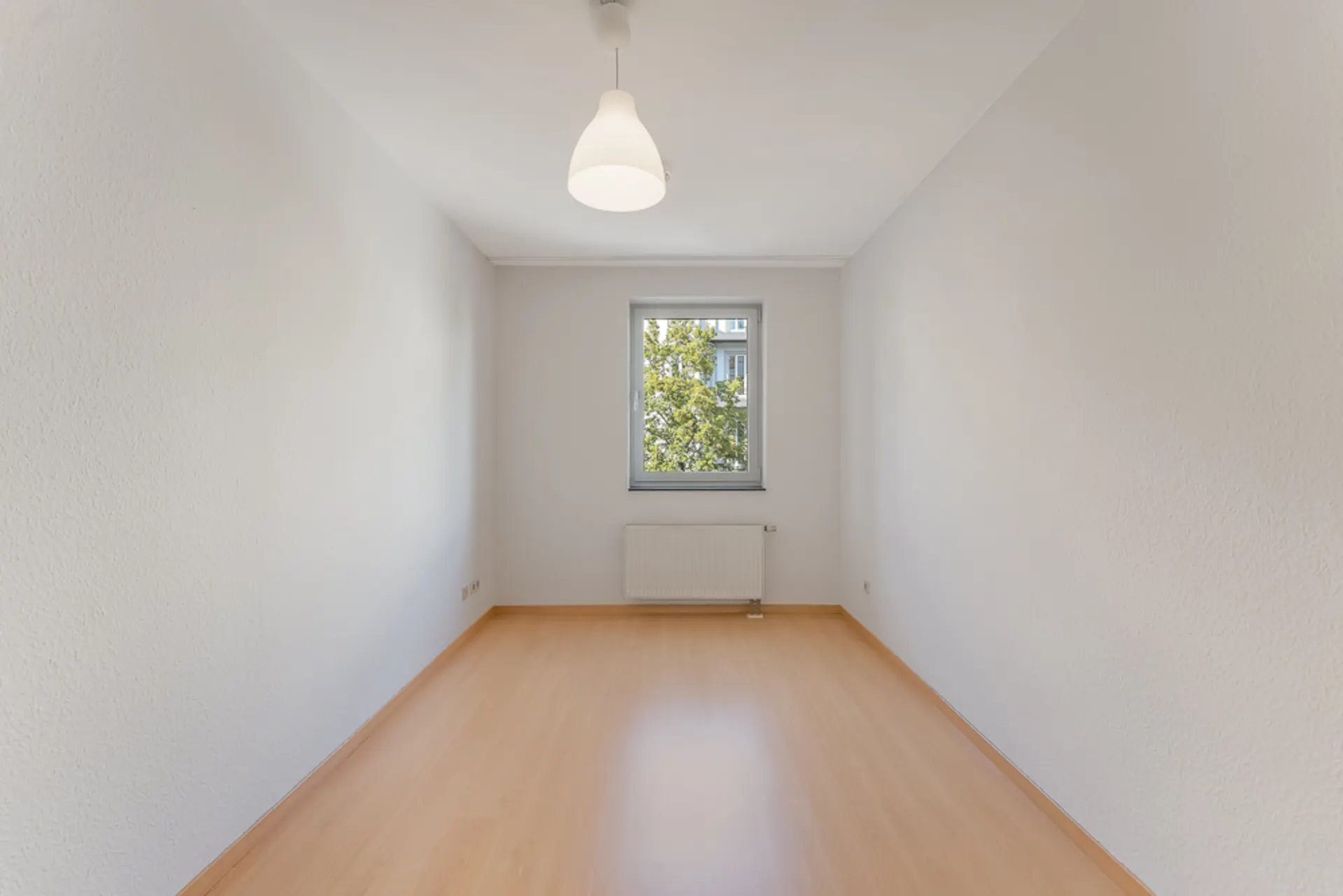 Leerer, heller Raum mit Holzboden, weißer Decke, Fenster, Heizung und Deckenlampe.