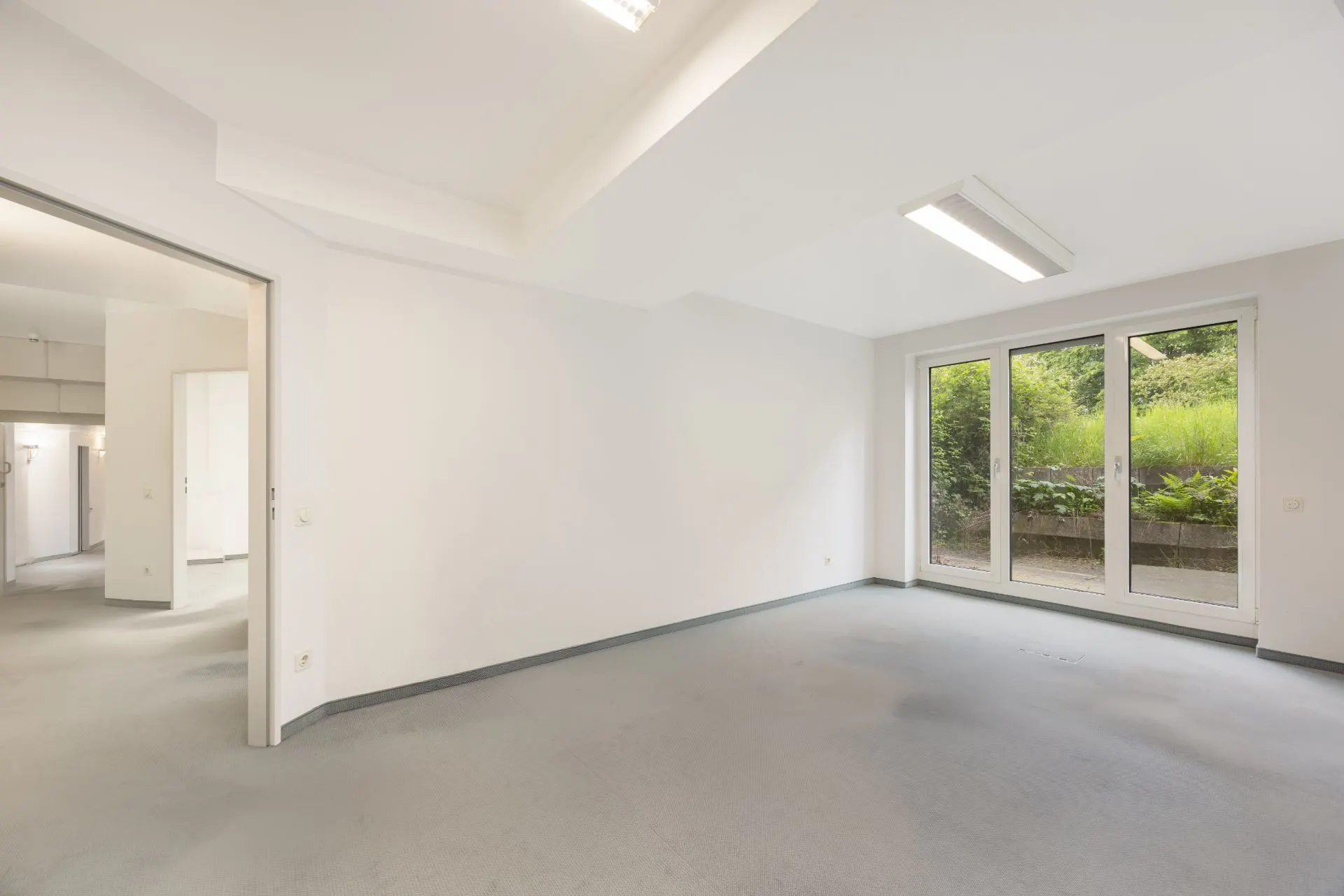 Heller leerer Raum mit großen Fenstern und Blick auf einen Garten.