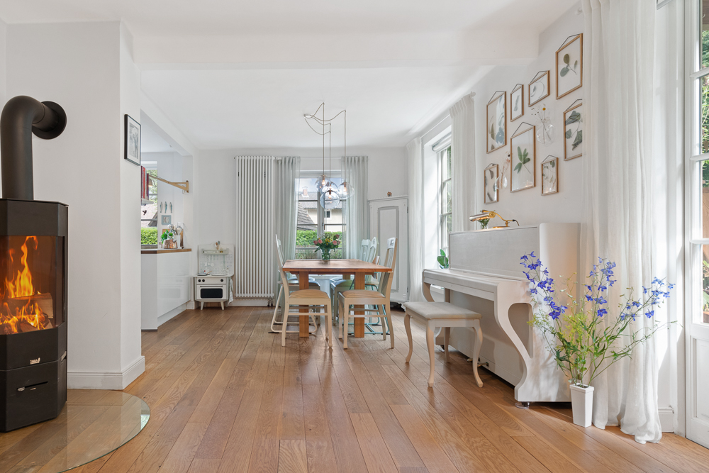 Helles Wohnzimmer mit Kamin, Holzboden, Esstisch, weißen Vorhängen und Klavier, Blumen und Wandbildern.