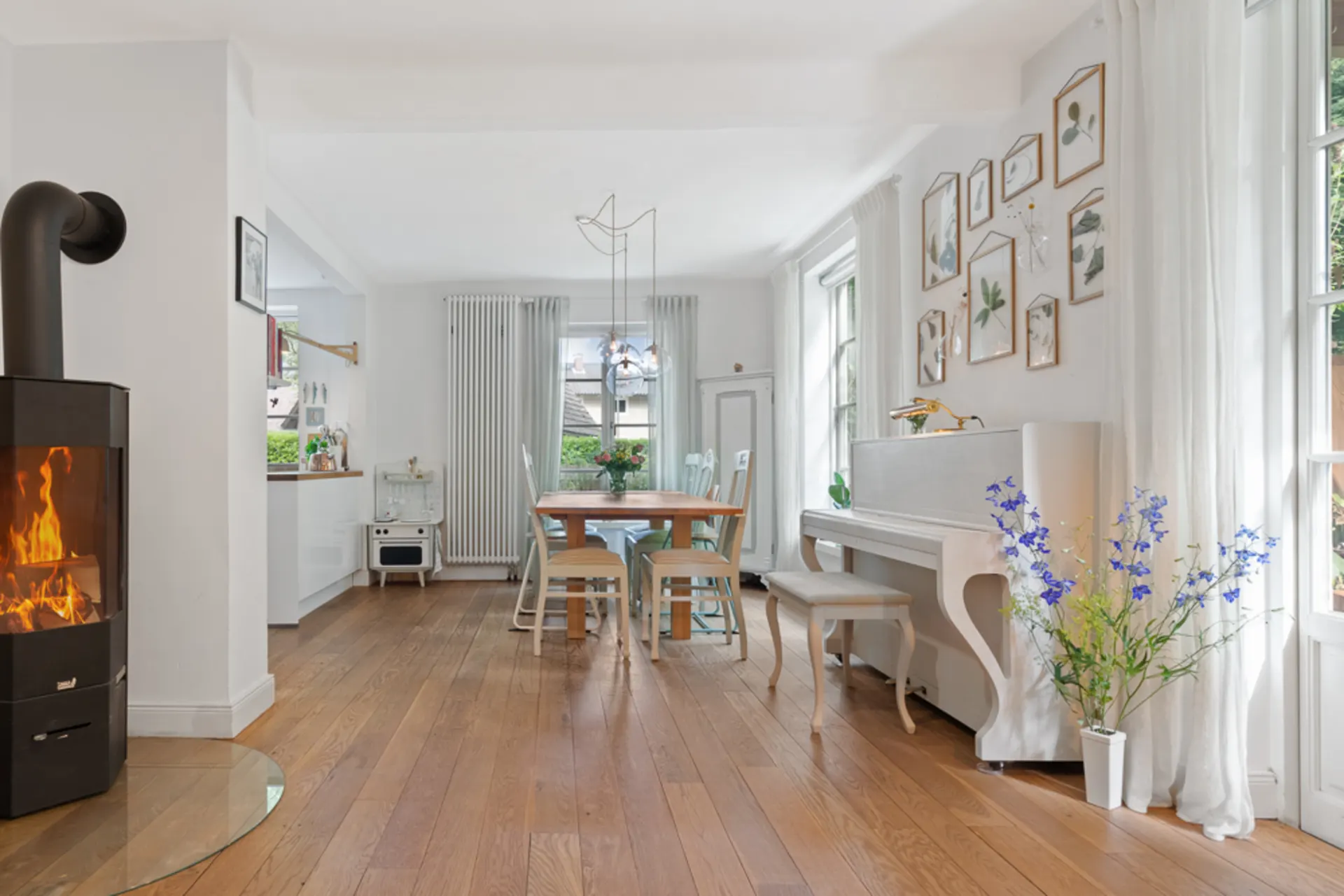 Helles Wohnzimmer mit Kamin, Holzboden, Esstisch, weißen Vorhängen und Klavier, Blumen und Wandbildern.
