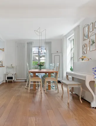 Helles Wohnzimmer mit Kamin, Holzboden, Esstisch, weißen Vorhängen und Klavier, Blumen und Wandbildern.