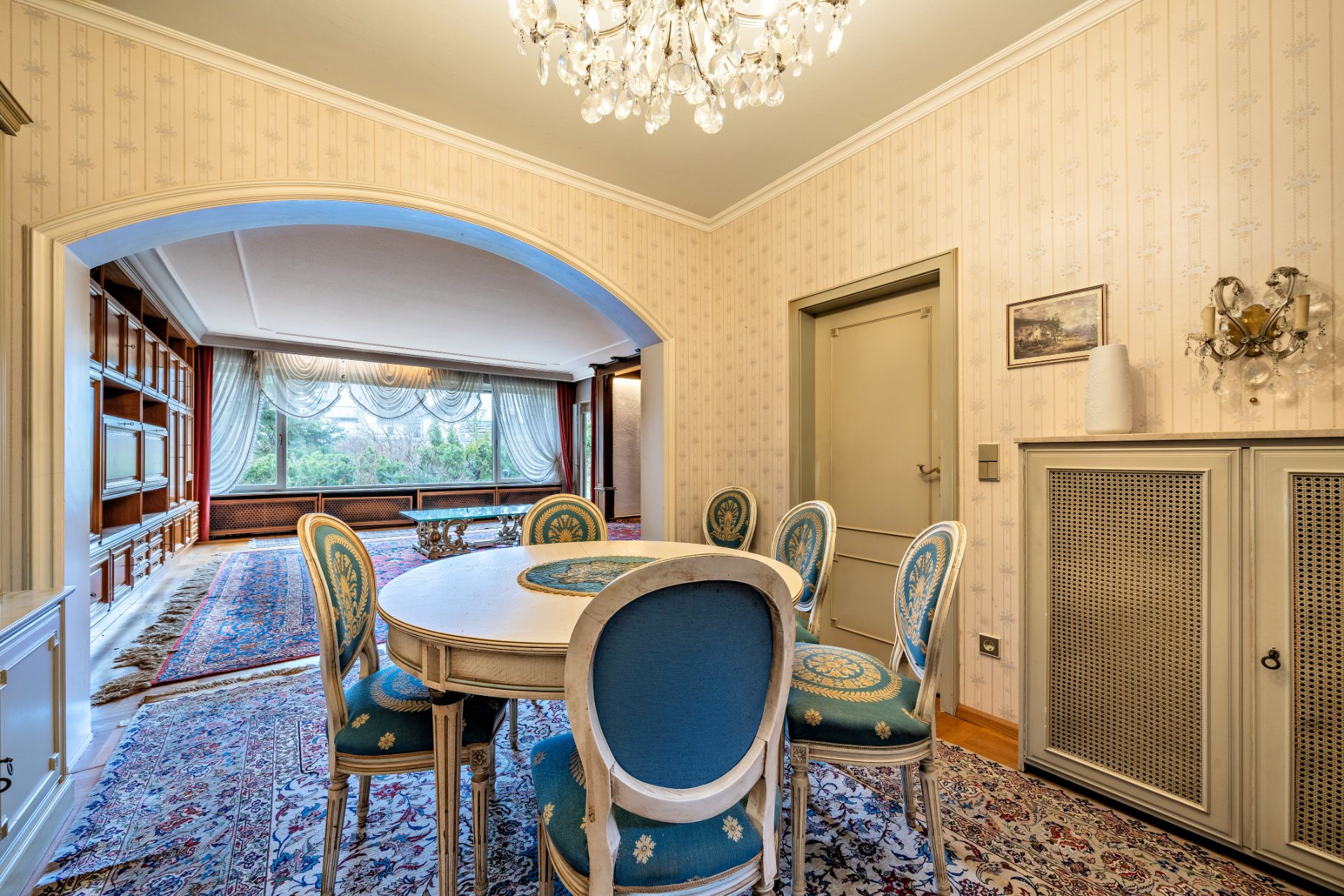 Esszimmer mit rundem Tisch und sechs Stühlen, Blick ins Wohnzimmer mit großen Fenstern.