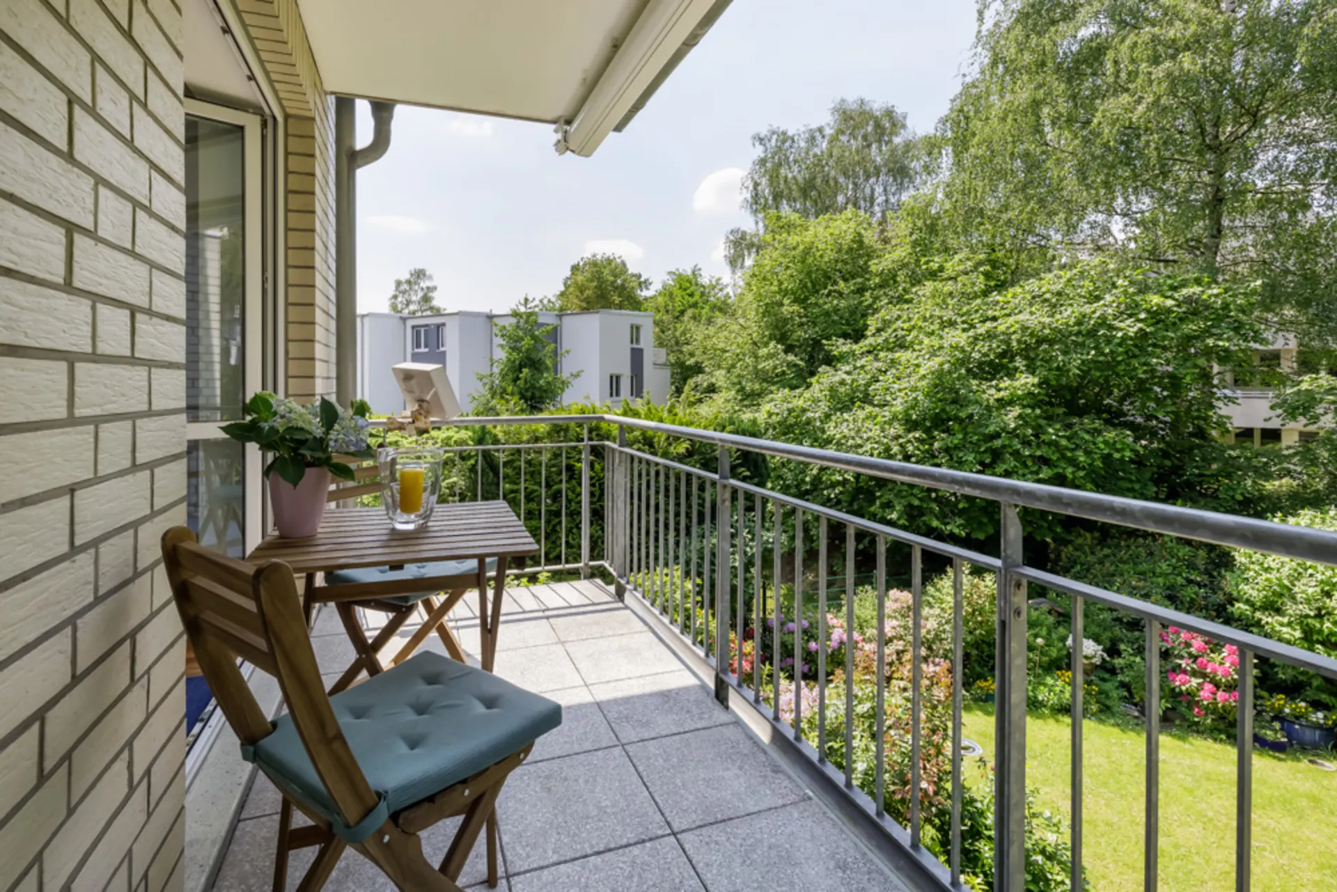 Balkon mit Holztisch, zwei Stühlen, Pflanze und Getränk, Blick auf grünen Garten.