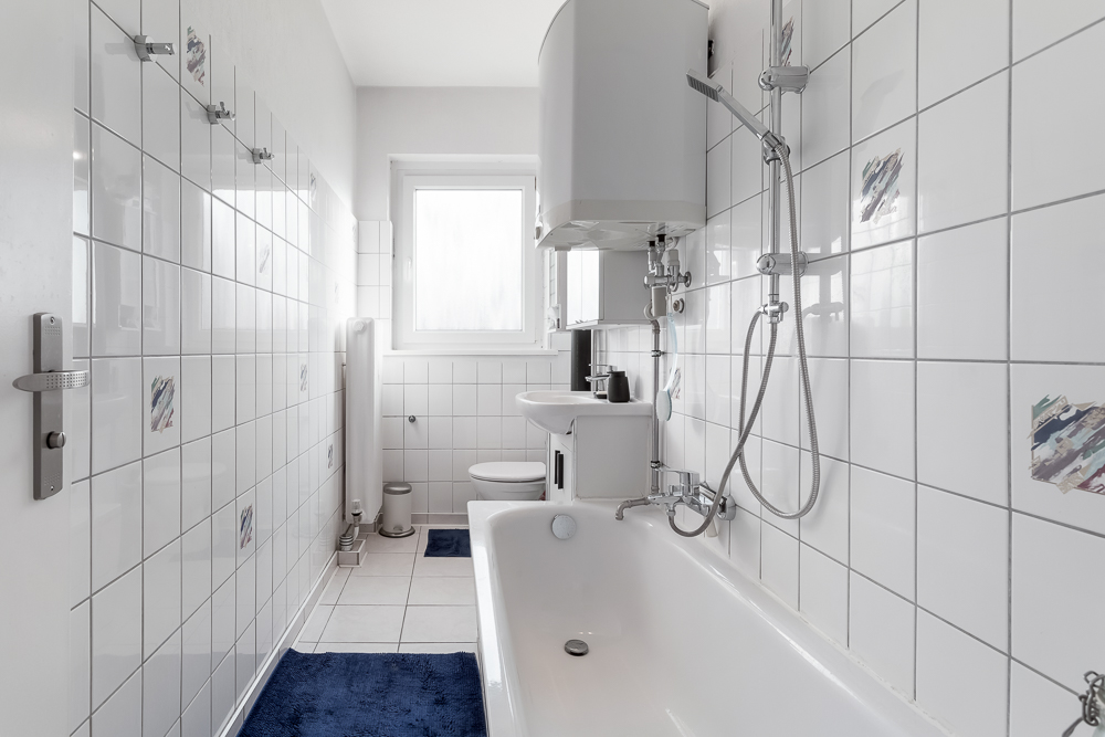 Schmales Badezimmer mit Badewanne, Dusche, Waschbecken und WC, weiße Fliesen und blauem Teppich.