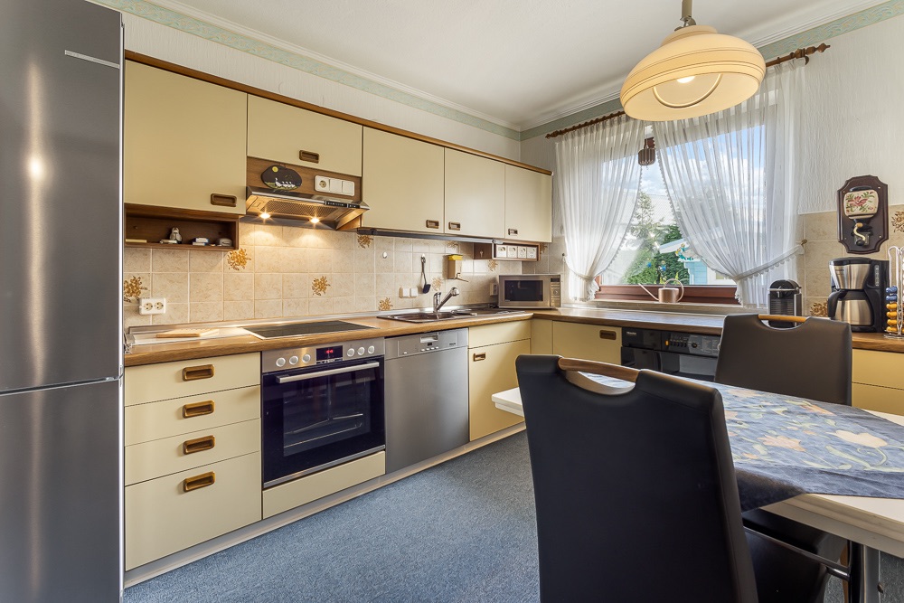 Helle Einbauküche mit Esstisch, Kühlschrank, Herd, Backofen und Kaffeemaschine, Fenster mit Vorhängen.