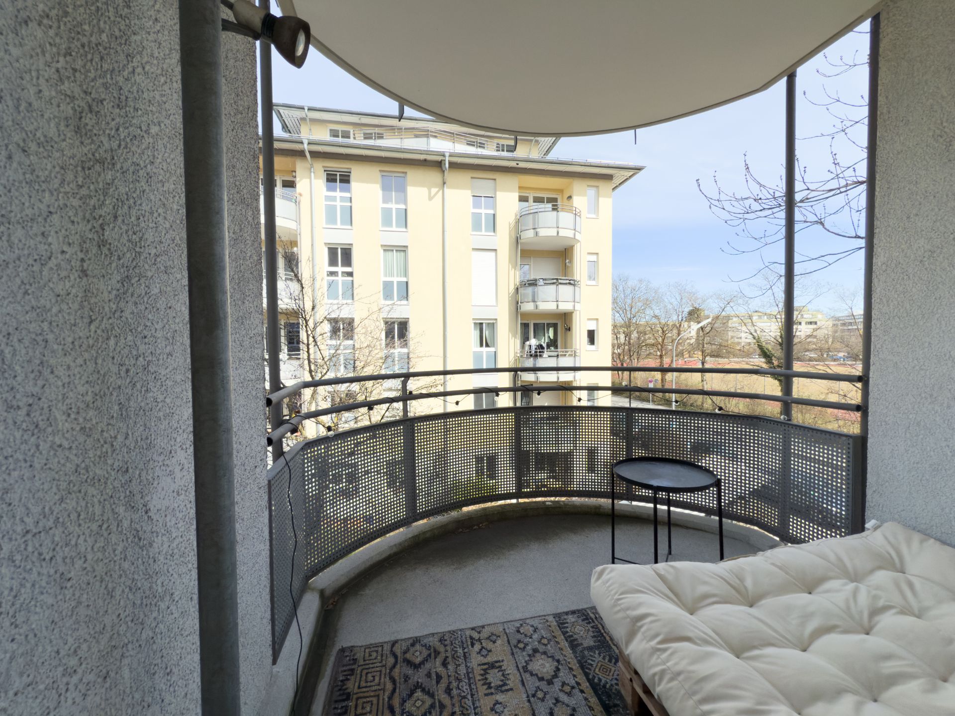 Überdachter Balkon mit Teppich, Tisch und Polsterbank, Blick auf Wohngebäude und Bäume.