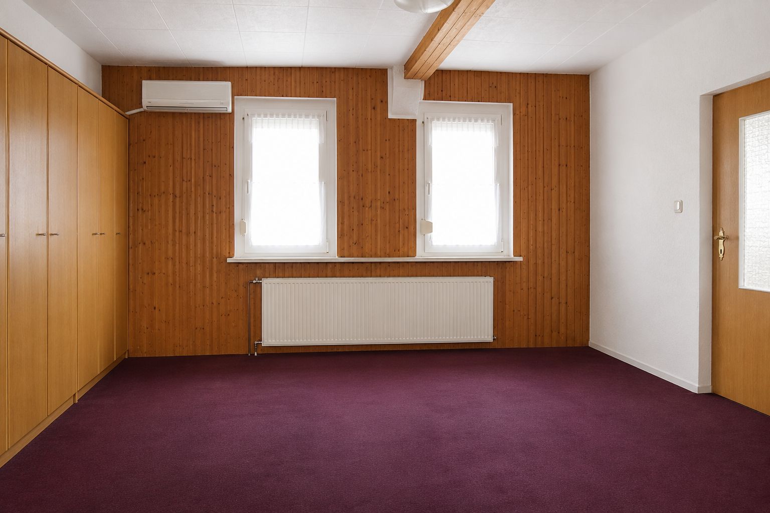 Leeres Zimmer mit Holzverkleidung, zwei Fenstern, Heizung, Einbauschrank und lila Teppichboden.