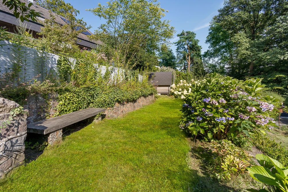 Grüner Garten mit Blumen, Holzbank an einer Steinmauer und Bäumen im Hintergrund.