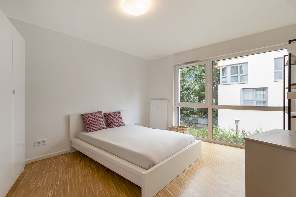 Helles Schlafzimmer mit großem Fenster, Doppelbett und zwei gemusterten Kissen.