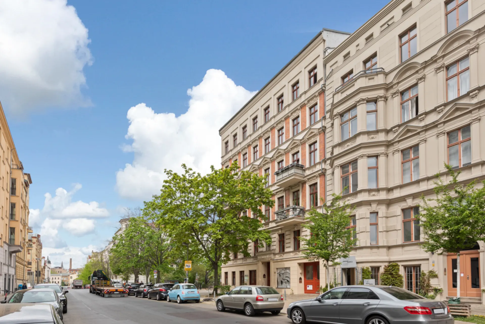 Wohnstraße mit mehrstöckigen Altbauten, Bäumen und parkenden Autos bei blauem Himmel.