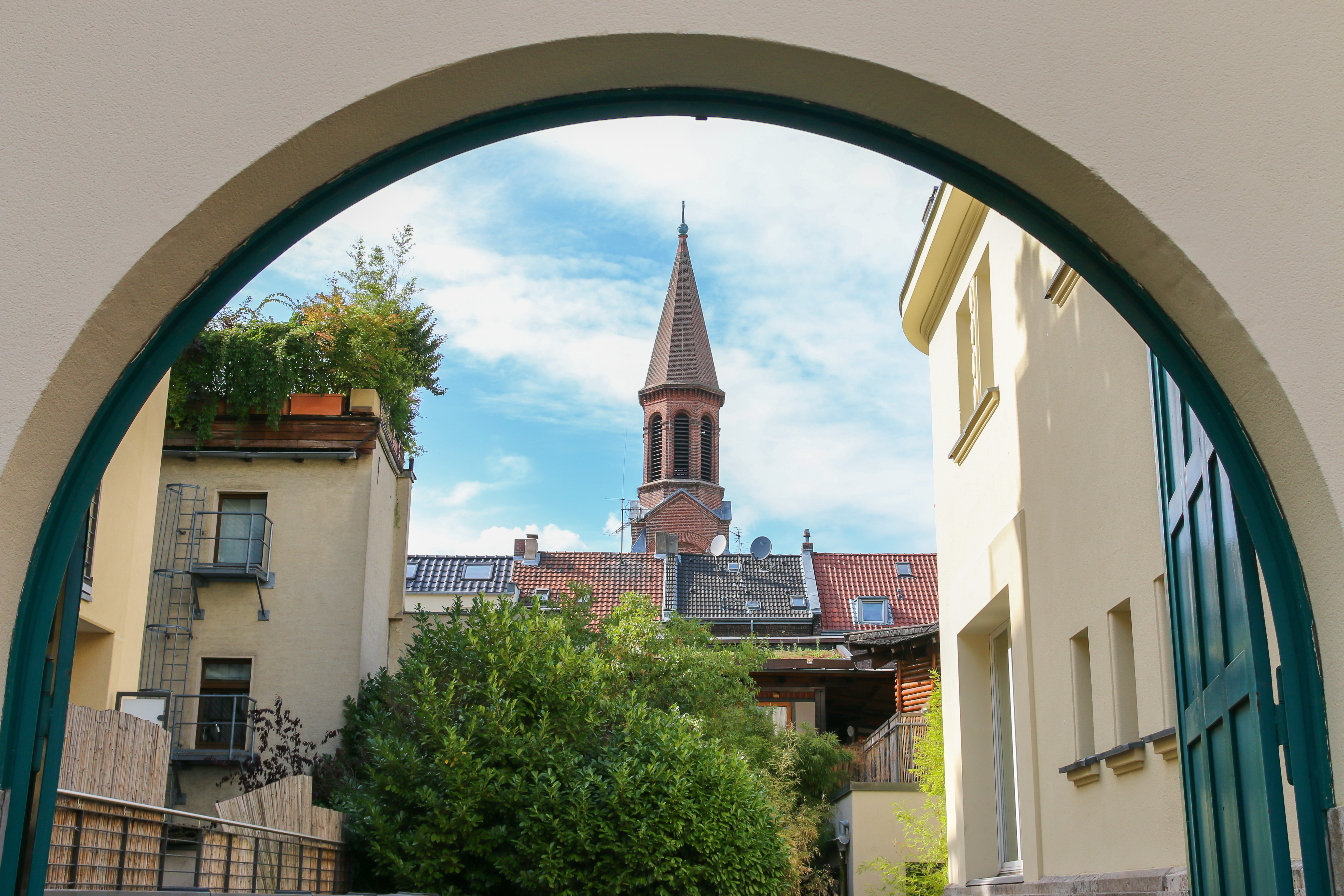 Helles Mehrfamilienhaus mit rotem Satteldach, vielen Fenstern und kleinem Erkerturm.