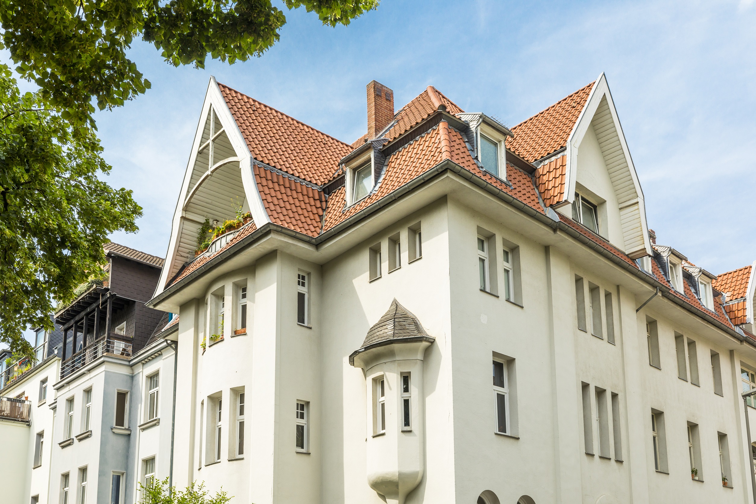 Helles Mehrfamilienhaus mit rotem Satteldach, vielen Fenstern und kleinem Erkerturm.