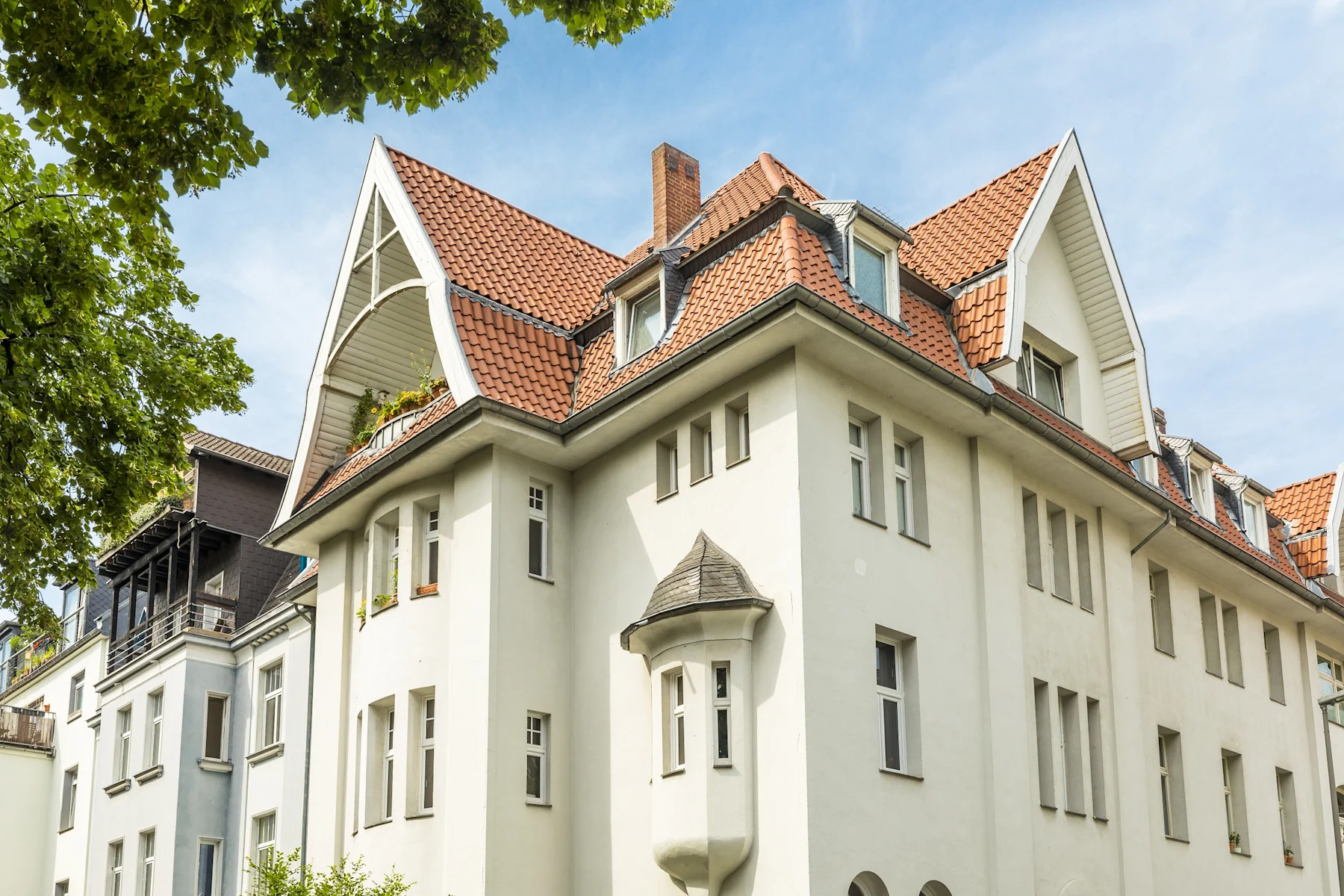 Helles Mehrfamilienhaus mit rotem Satteldach, vielen Fenstern und kleinem Erkerturm.