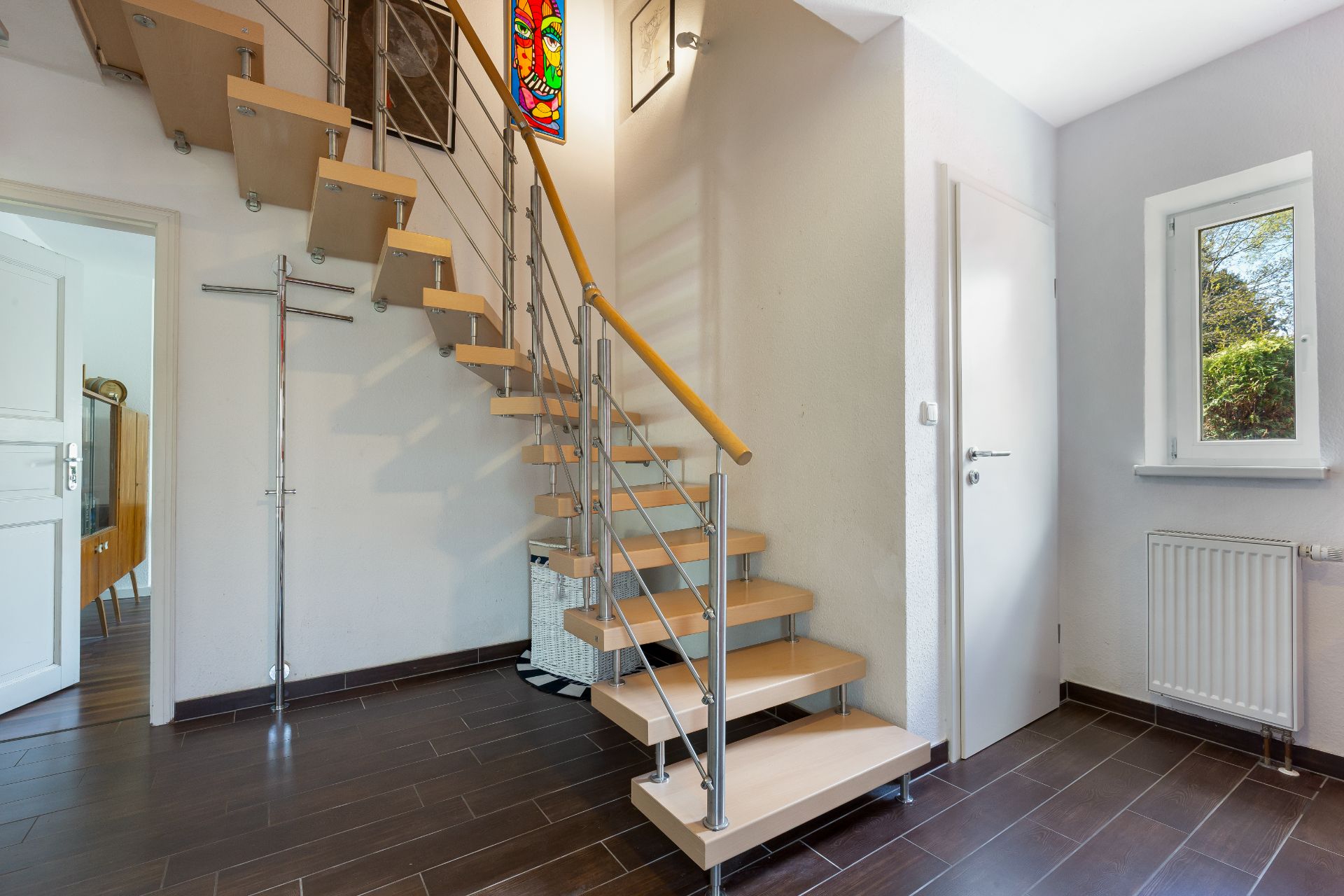 Moderne Holztreppe mit Metallgeländer in einem hellen Flur mit braunem Fliesenboden.