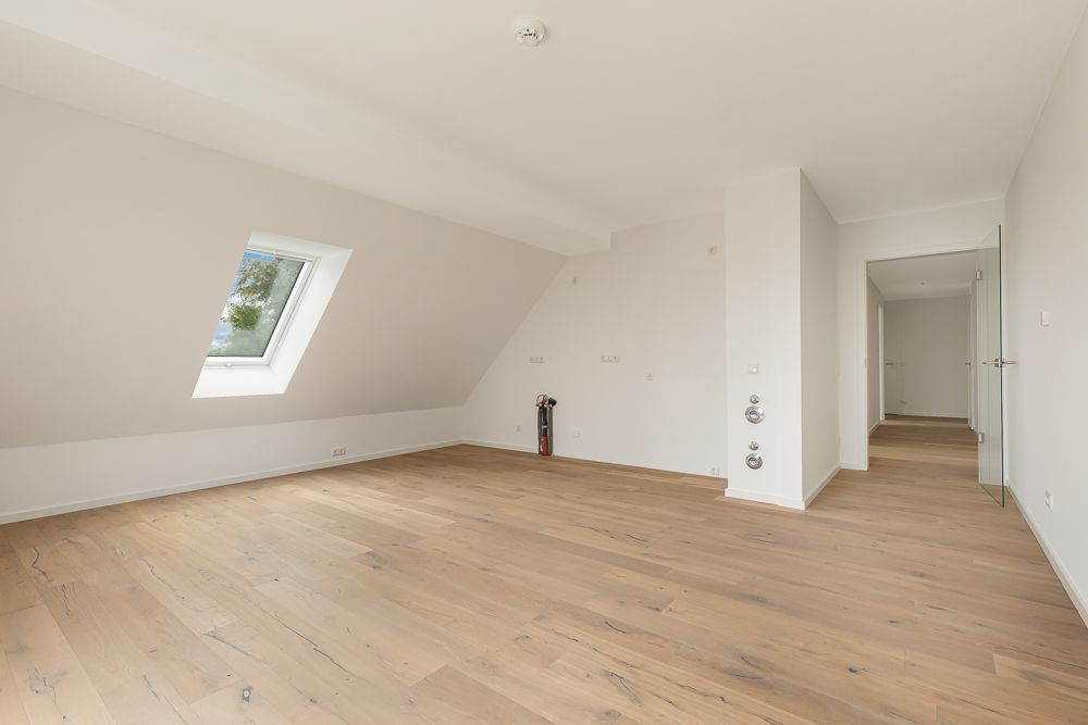 Helles, leeres Zimmer mit Holzfußboden, Dachschrägen und einem Fenster auf der linken Seite.