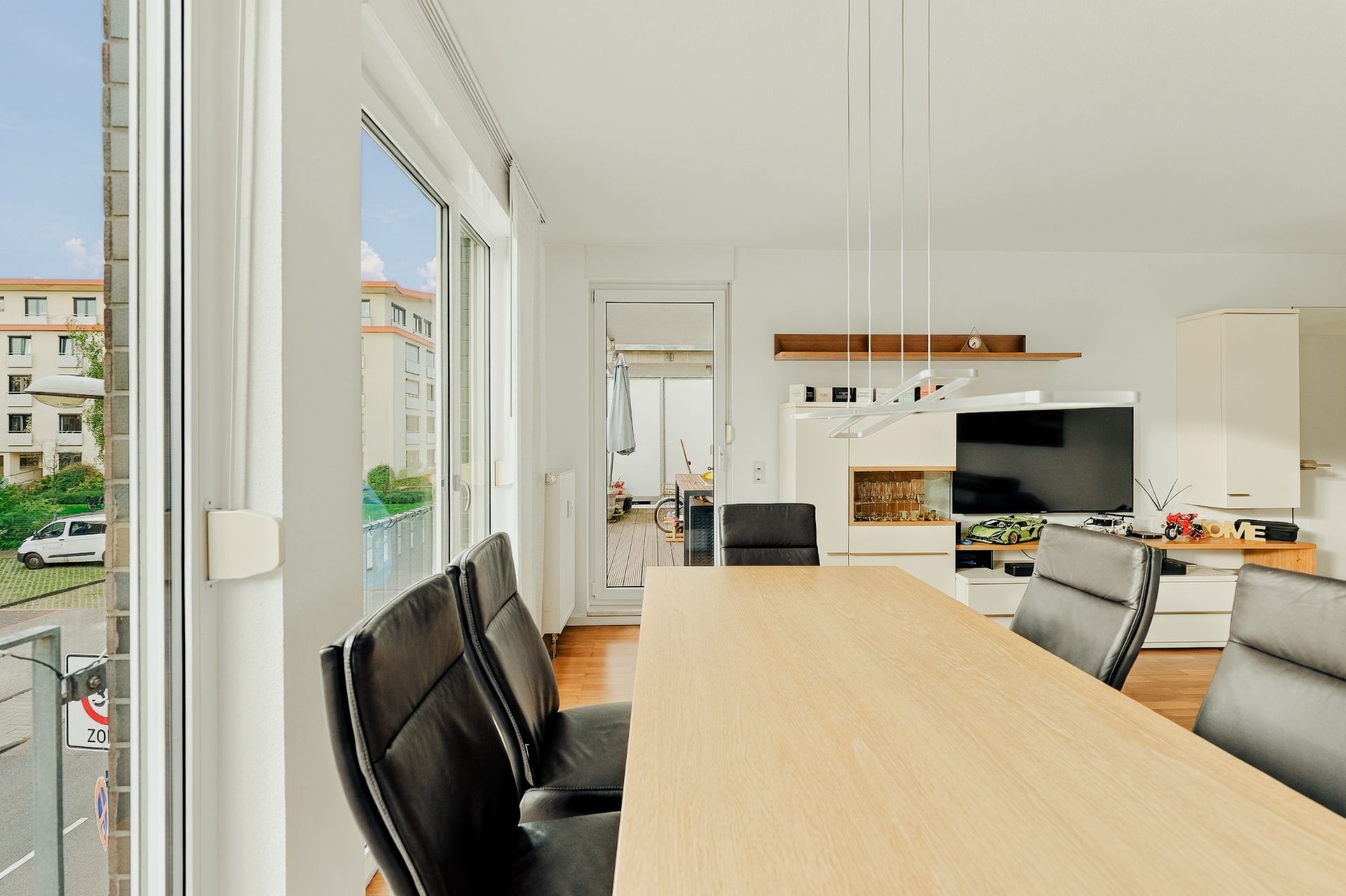 Essbereich mit Holztisch und schwarzen Stühlen, große Fenster und Blick ins Wohnzimmer mit Fernseher.
