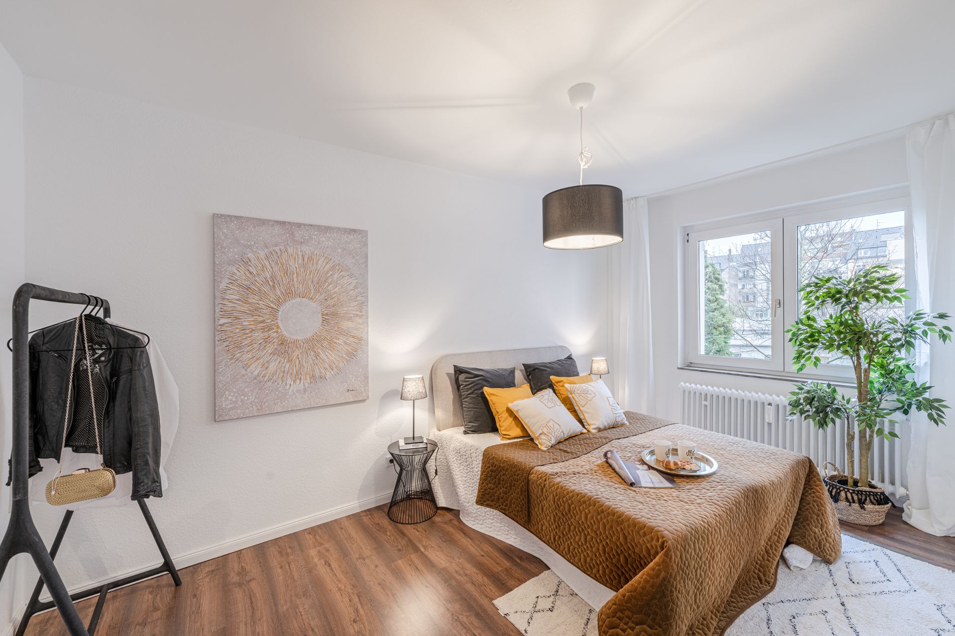 Helles Schlafzimmer mit Doppelbett, Bildern an der Wand, Kleiderständer und großem Fenster.