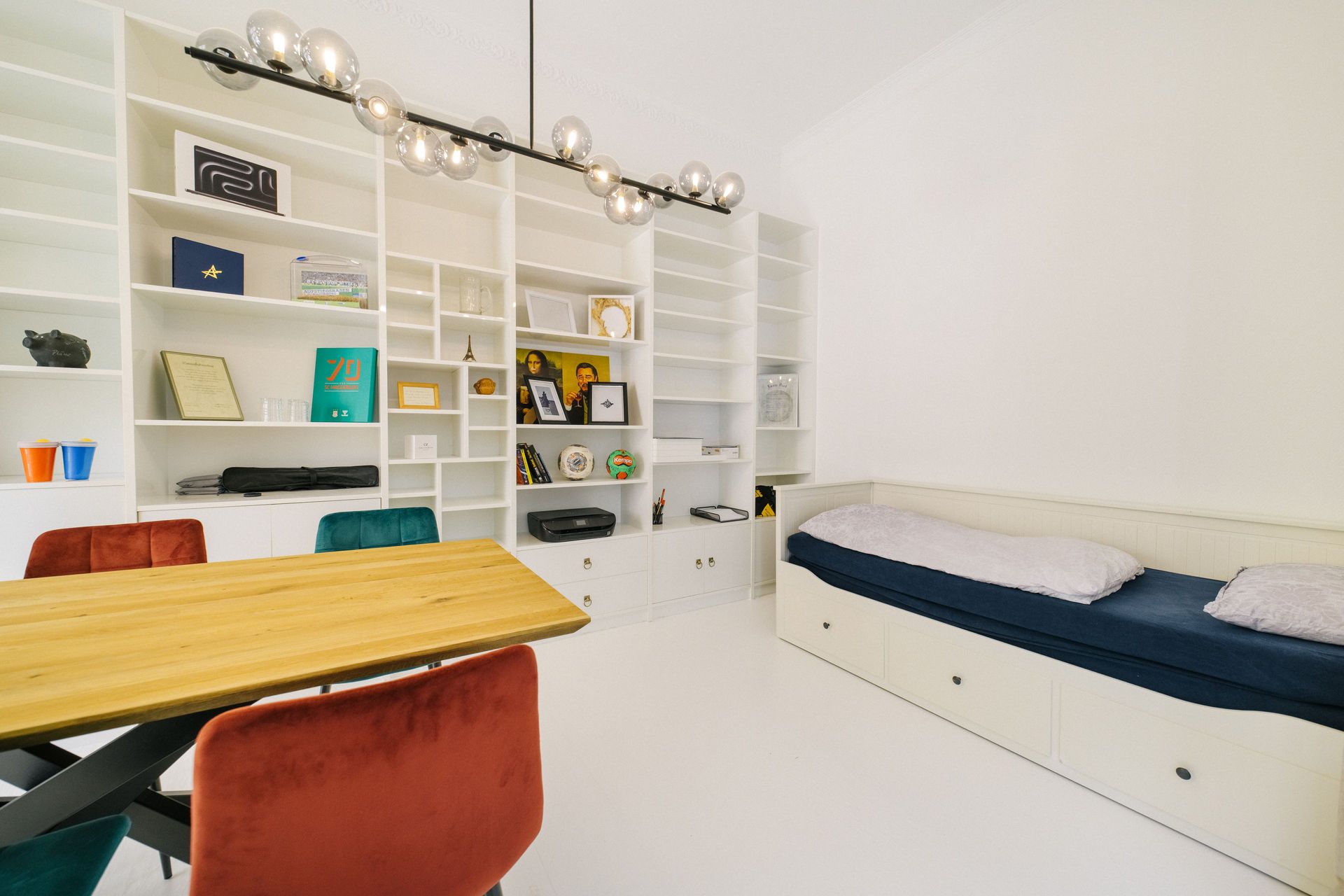 Helles Zimmer mit Bücherregal, Holztisch, bunten Stühlen und Tagesbett mit blauer Decke.