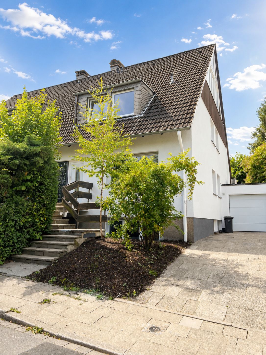 Doppelhaushälfte mit Vorgarten, Treppe zum Eingang und Auffahrt zur Garage bei sonnigem Wetter.