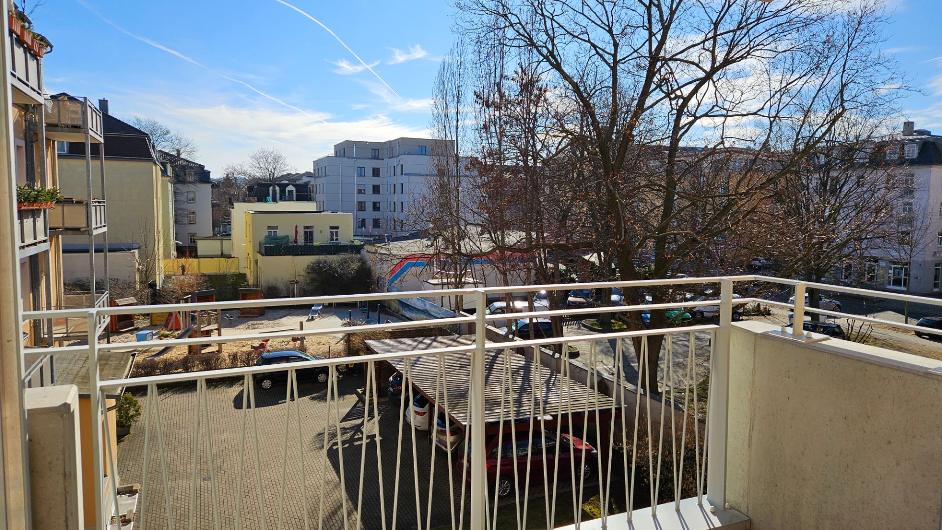 Blick von einem Balkon auf einen Innenhof mit Bäumen, Autos und umliegenden Wohngebäuden.