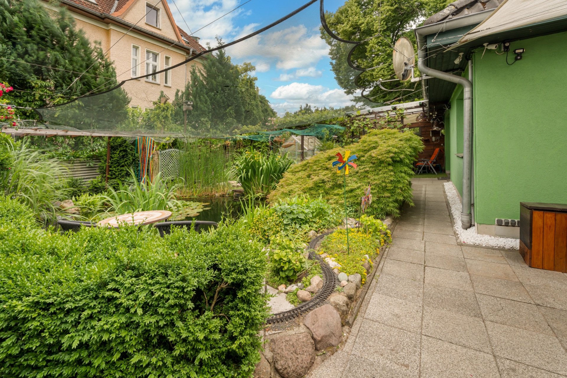 Gepflegter Garten mit Teich, Sträuchern, Gehwegplatten und einem Spielzeug-Windrad neben einem grünen Haus.
