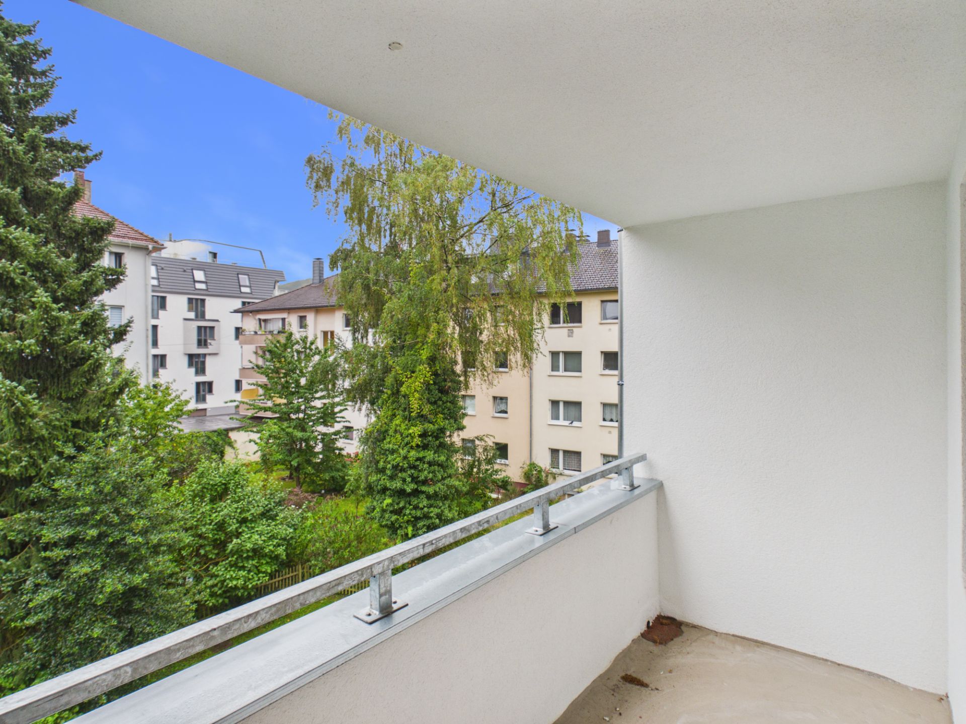 Überdachter Balkon mit Blick auf grüne Bäume und benachbarte Wohngebäude.