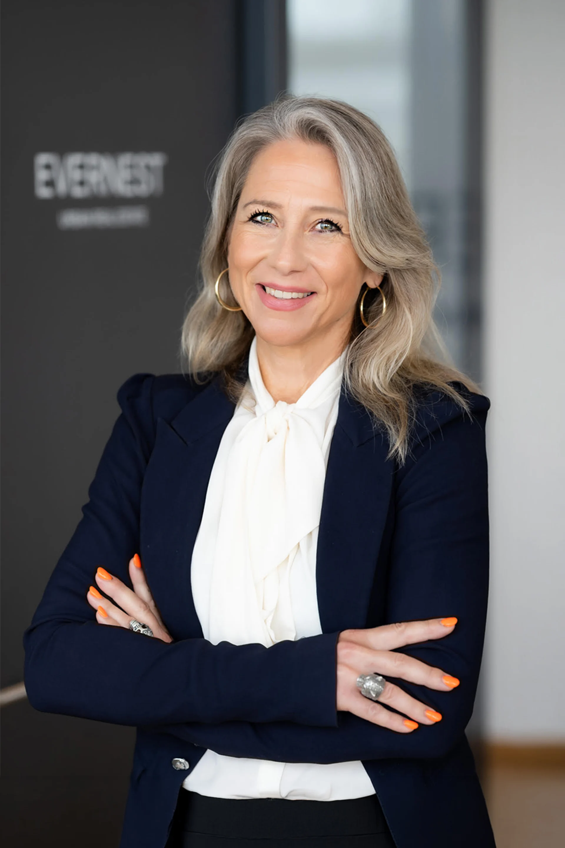 Frau mit blonden Haaren im Business-Outfit steht mit verschränkten Armen und lächelt.