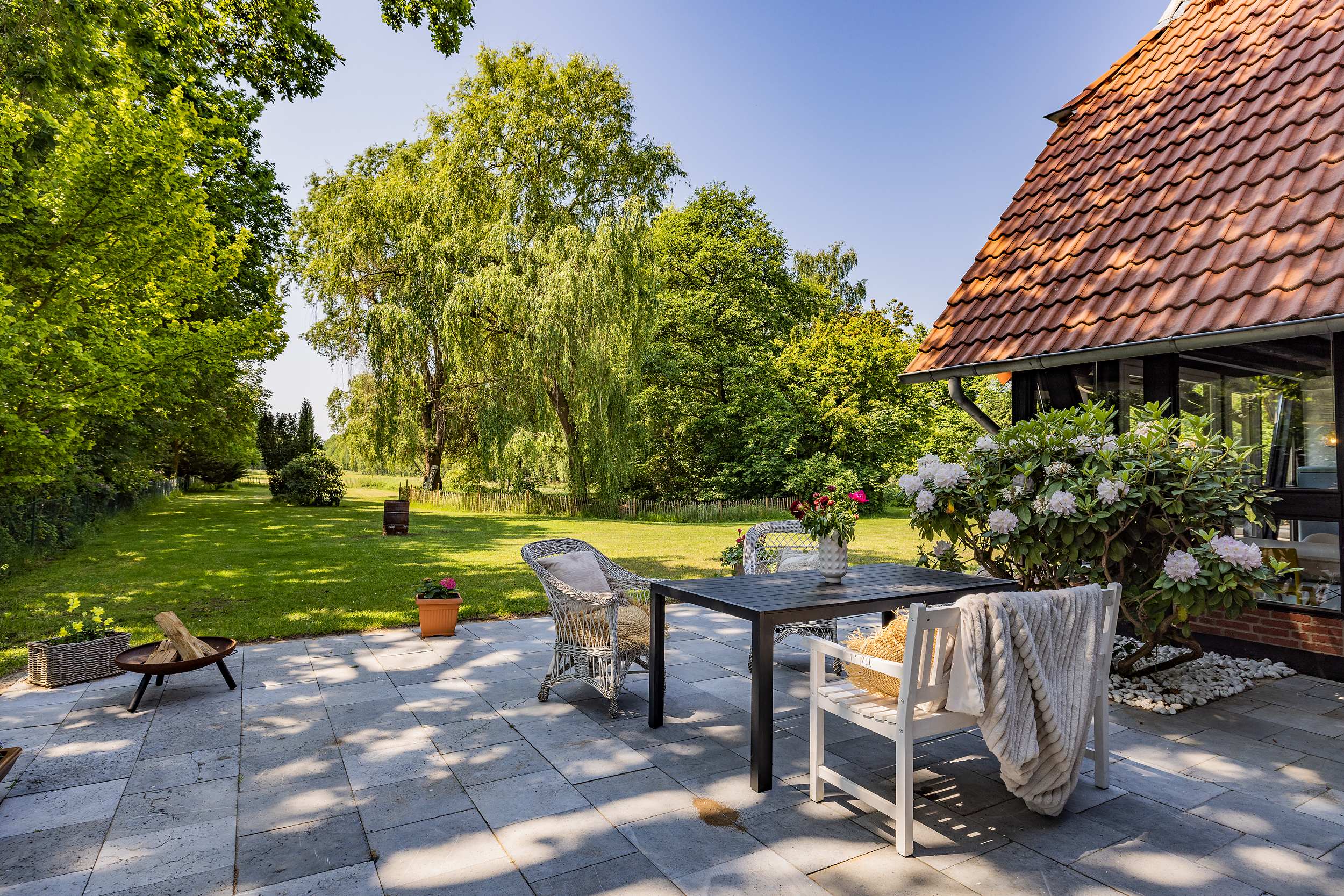 Gepflasterte Terrasse mit Sitzgruppe und Blumen, daneben großer grüner Garten mit Bäumen.
