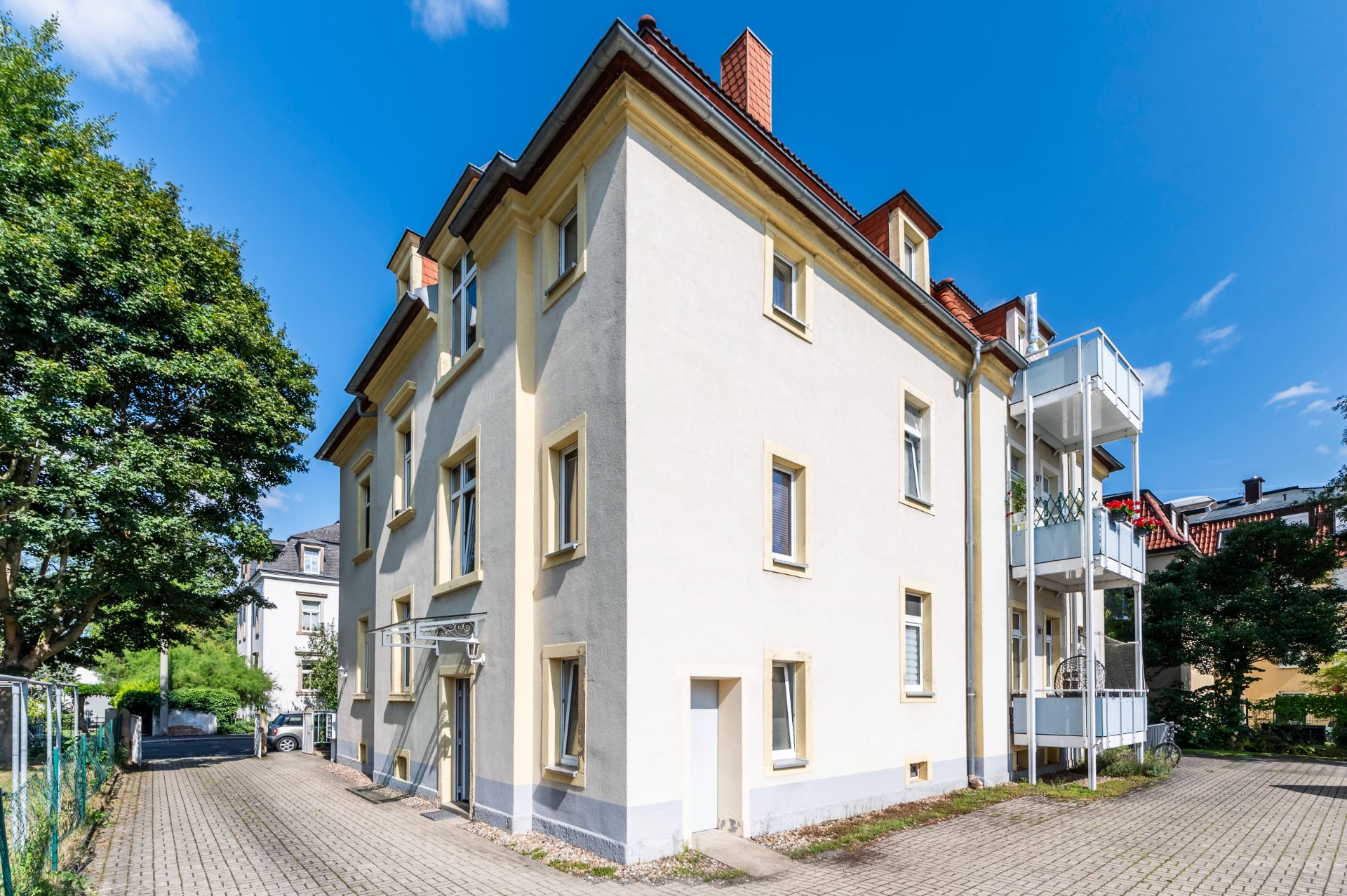 Helles, dreistöckiges Wohnhaus mit Satteldach und kleinem Balkon, umgeben von Bäumen und Pflasterweg.