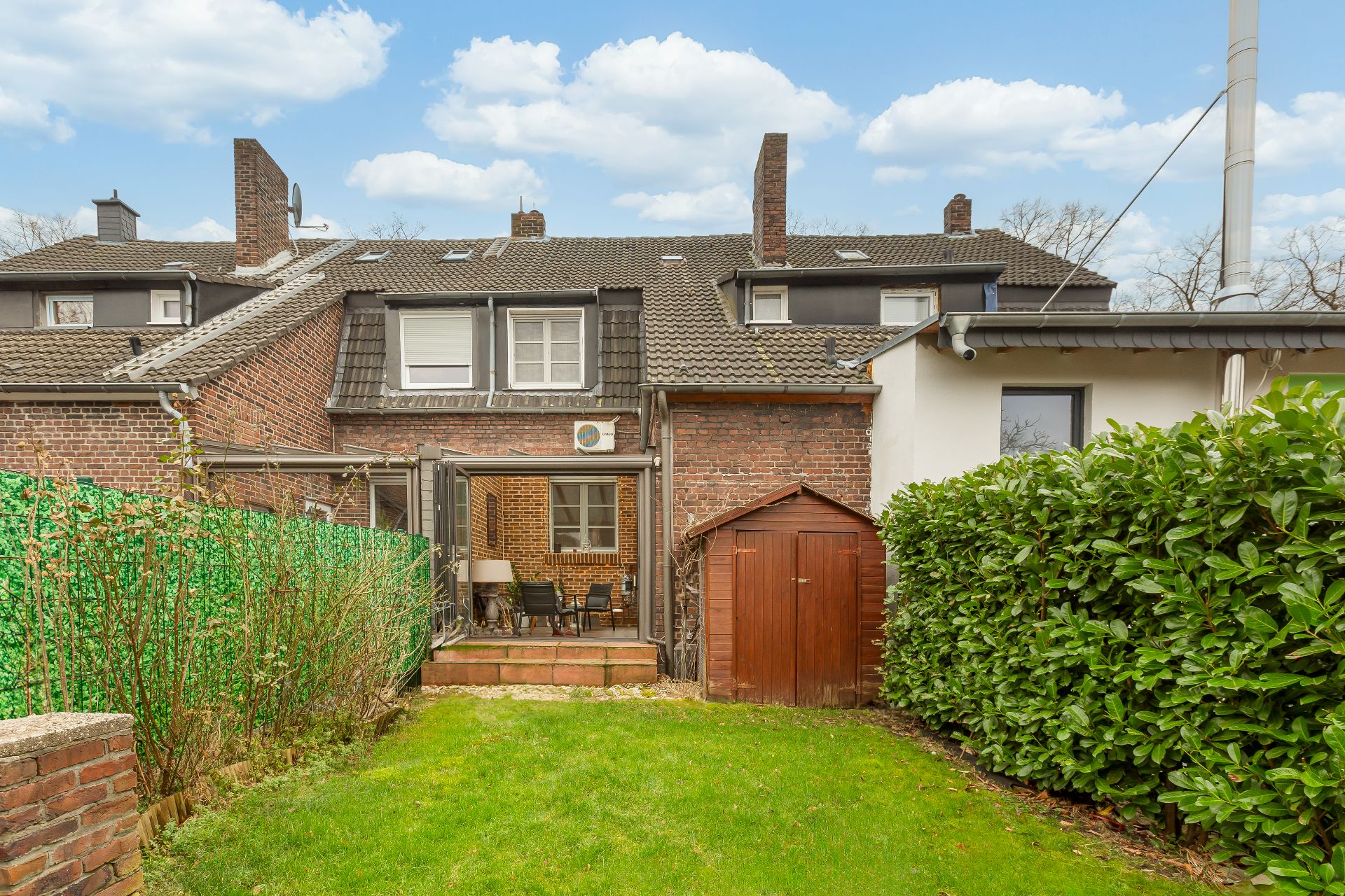 Rückseite eines Backsteinhauses mit Terrasse, Garten und Holzschuppen.