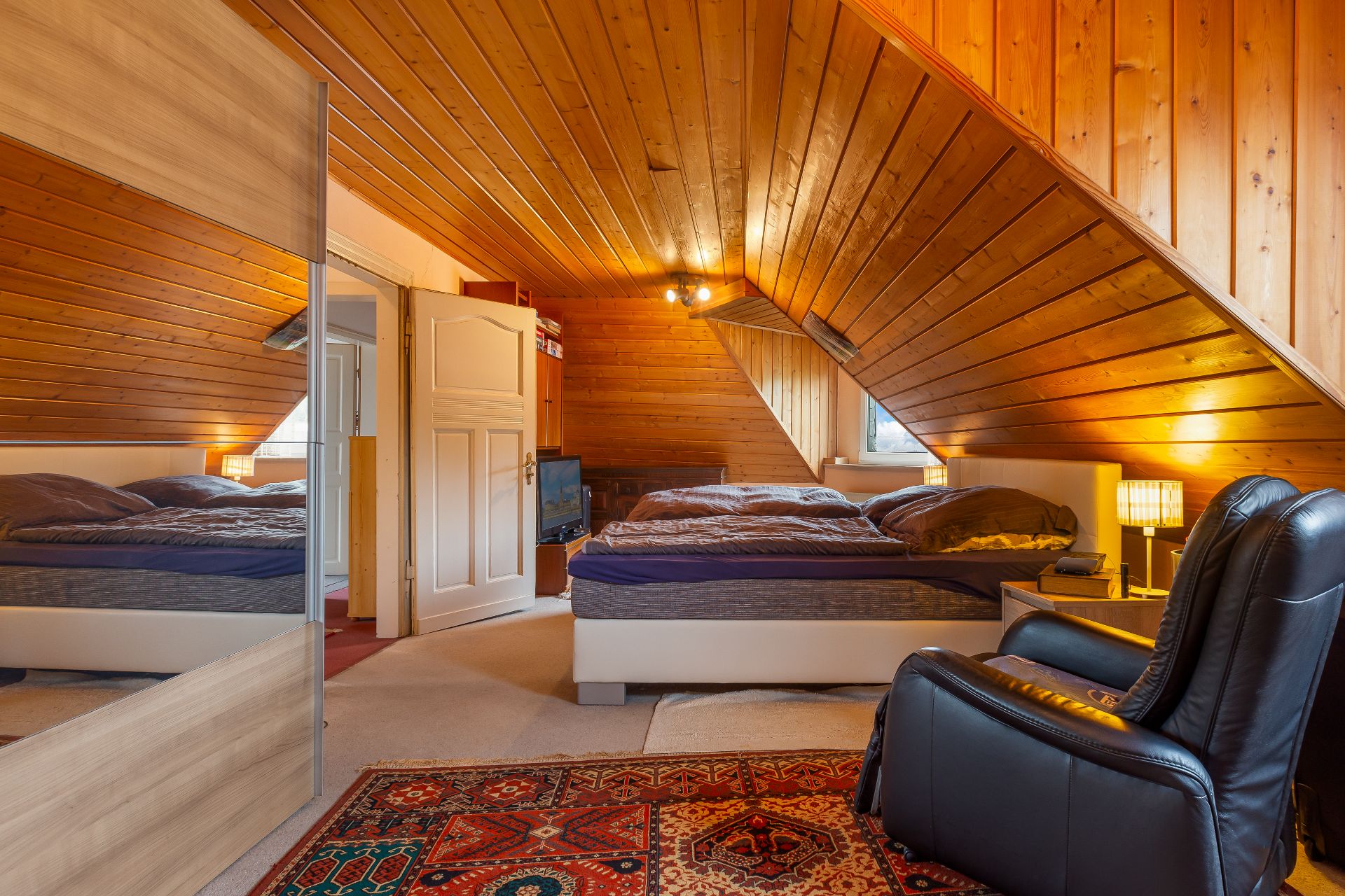 Dachzimmer mit Holzdecke, Doppelbett, Sessel, Teppich und Nachttischen mit Lampen.