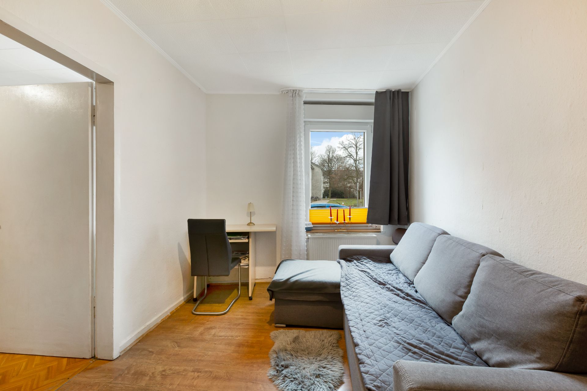 Wohnzimmer mit grauem Sofa, kleinem Schreibtisch und Stuhl vor einem Fenster mit Vorhängen.
