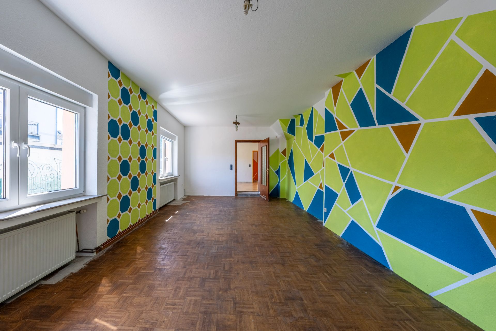 Heller Raum mit Parkettboden und bunt gemusterten Wänden, große Fenster auf der linken Seite.