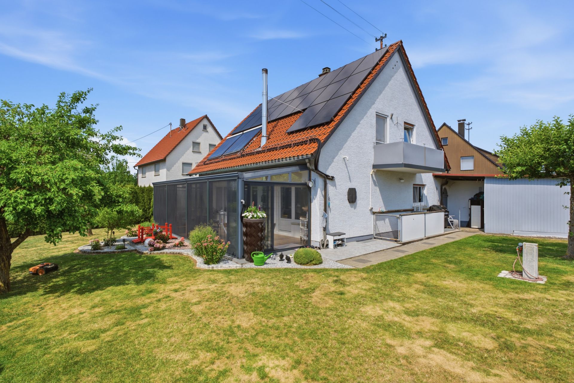 Weißes Einfamilienhaus mit Solarpanelen, Wintergarten und großem Garten mit Bäumen.