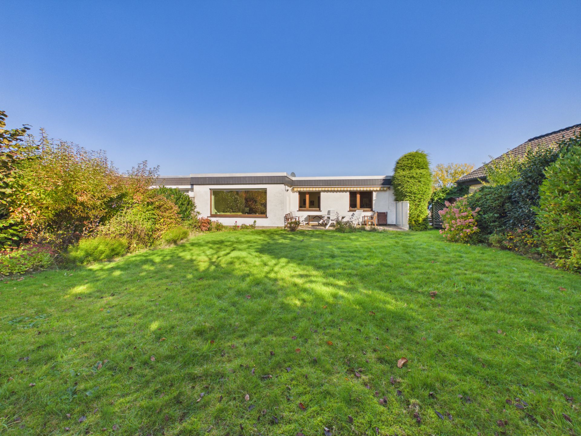 Einfamilienhaus mit großer Terrasse und grünem Garten, umgeben von Büschen und Bäumen.